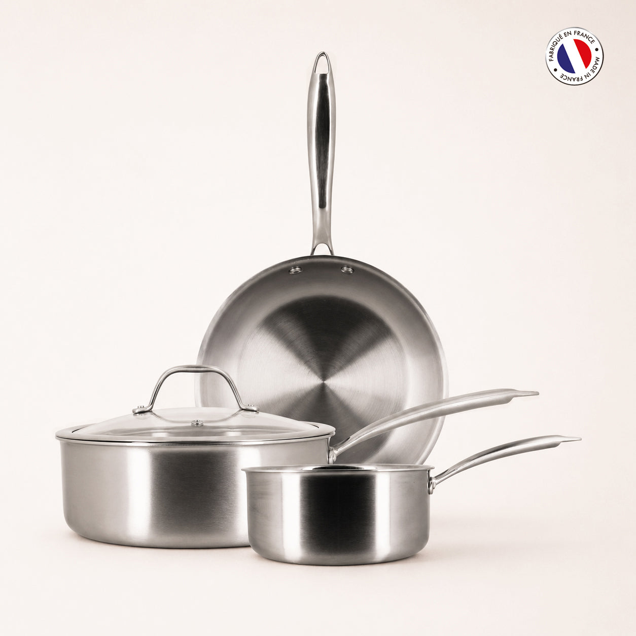 Set de 3 pièces Triply Menastyl en acier inoxydable : poêle 28cm, casserole 18cm et sauteuse 24cm empilées sur fond beige