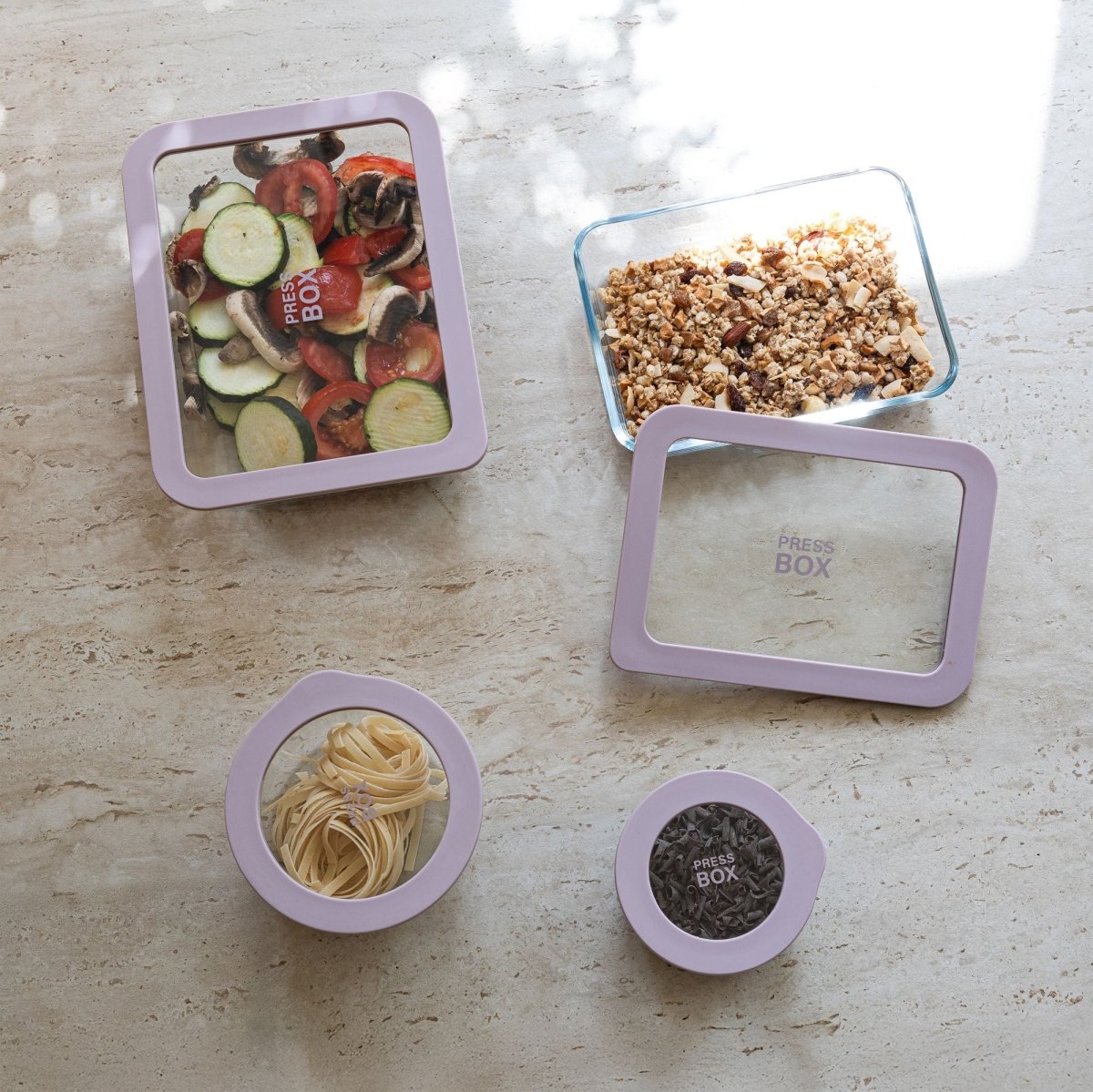 Ensemble de boîtes hermétiques Pressbox en verre avec couvercles violets contenant différents aliments sur plan de travail clair
