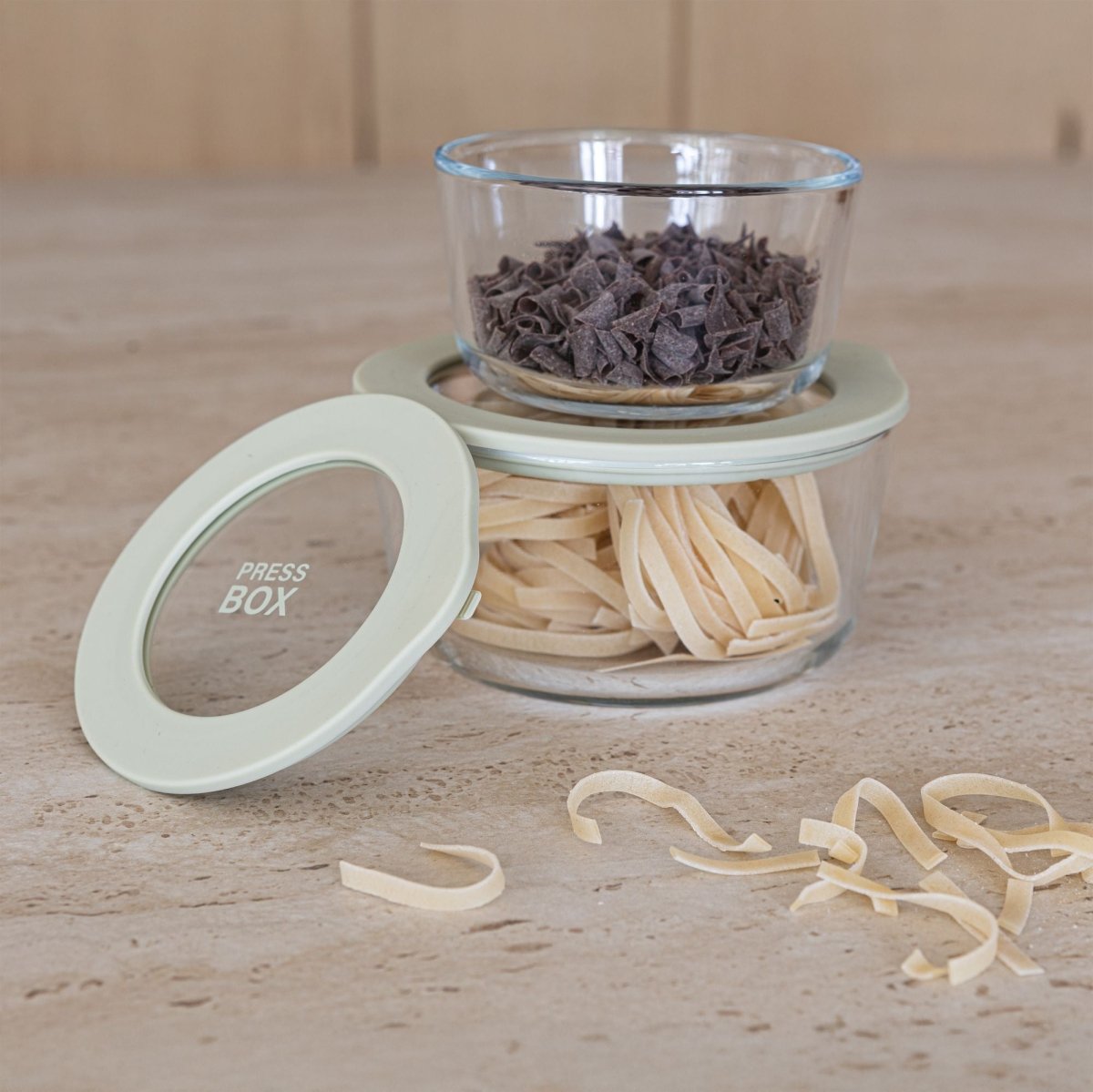 Boîte hermétique ronde Pressbox en verre borosilicate avec pâtes fraîches et pépites de chocolat superposées sur plan de travail