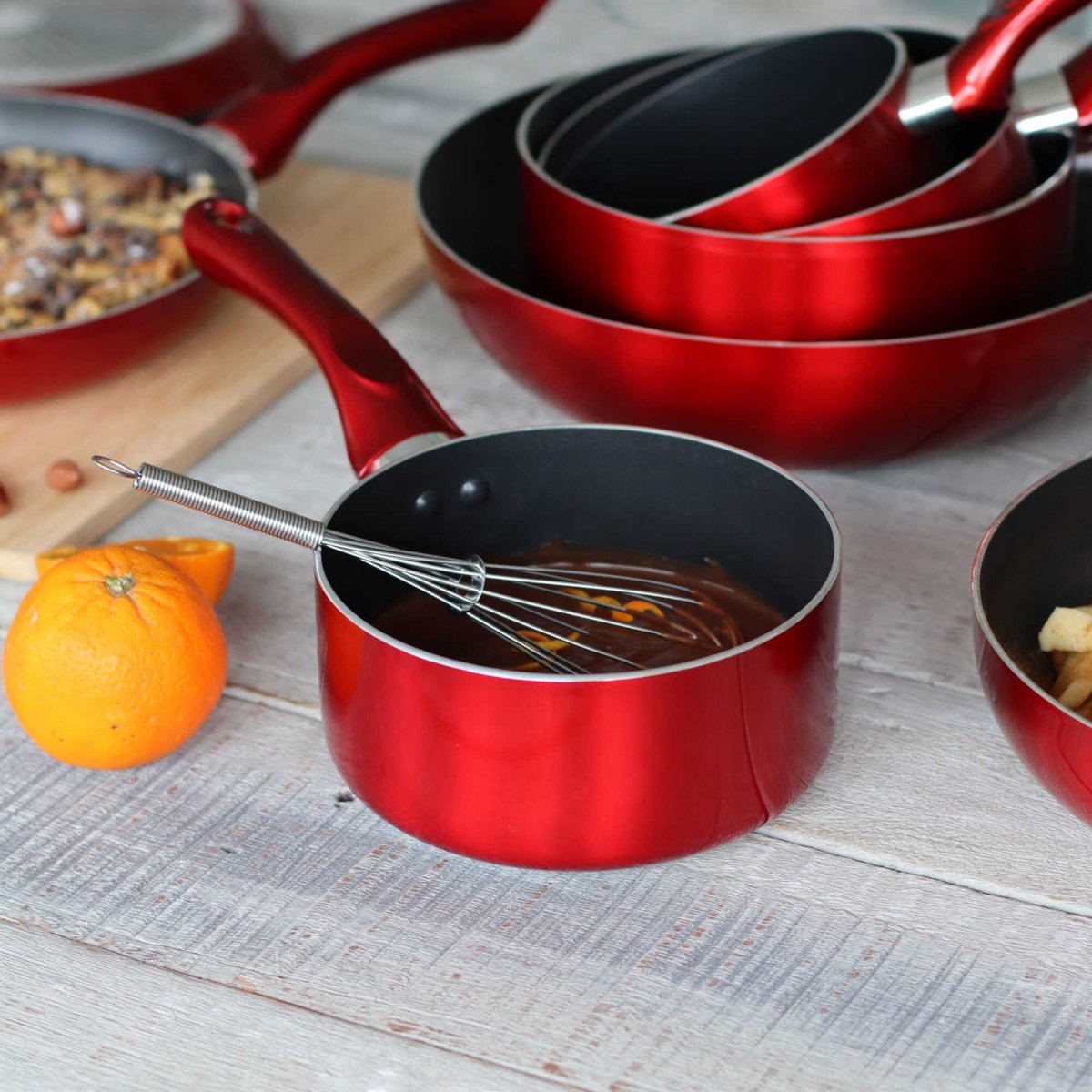 Set de casseroles Cerise rouges avec intérieur antiadhésif noir, whisk et orange, disposition lifestyle sur table bois