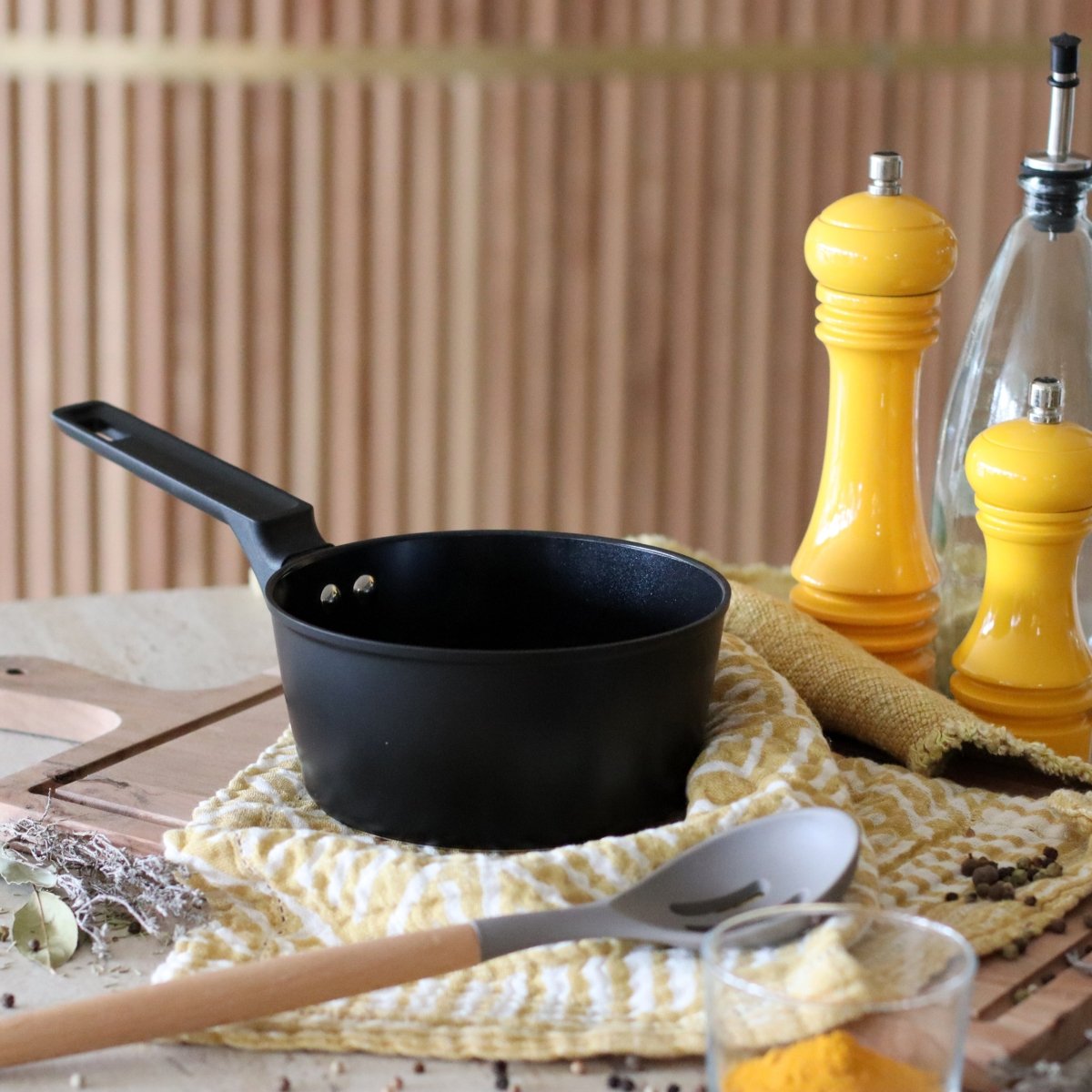 Casserole First 16 cm en aluminium noir sur table avec moulin à poivre jaune et condiments