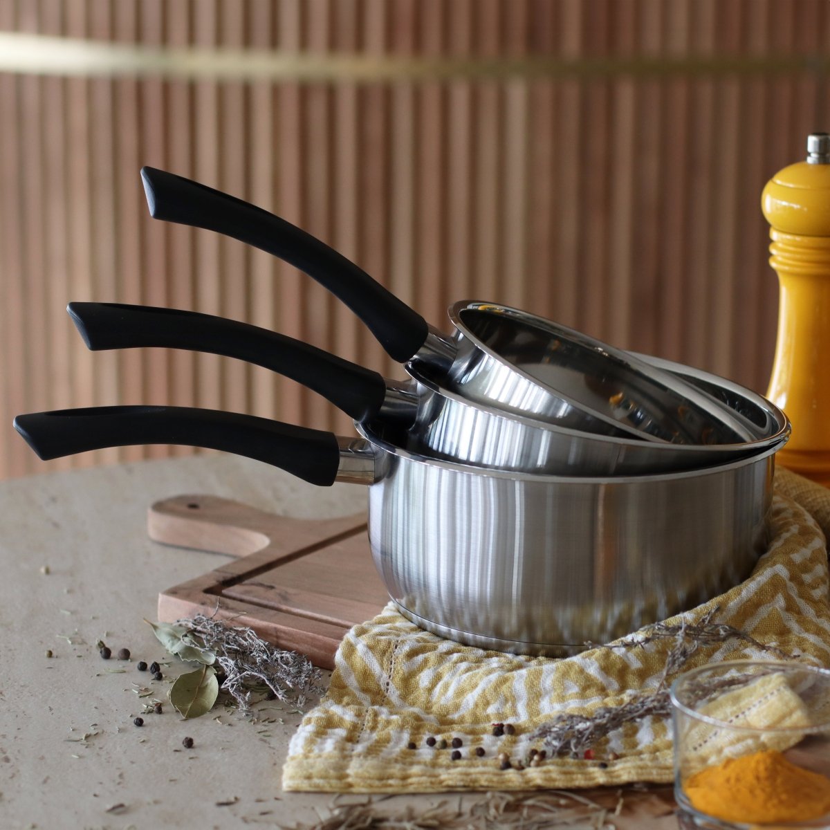 Set de 3 casseroles First en acier inoxydable avec couvercle et poignées noires, sur plan de travail avec textile jaune