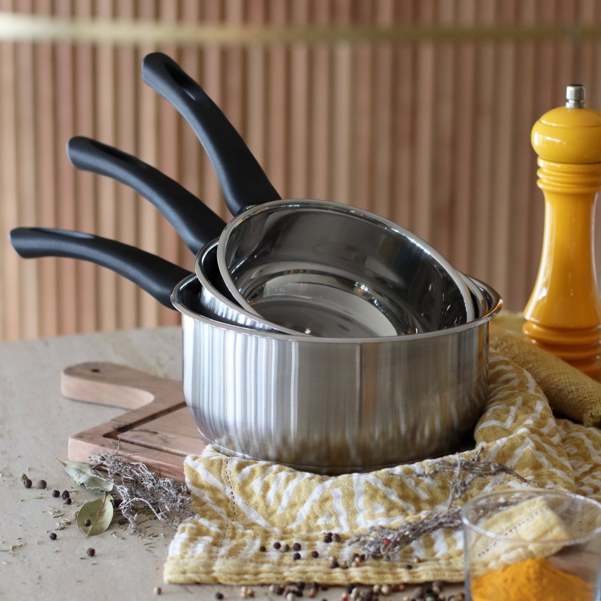 Set de 3 casseroles First en acier inoxydable avec manches noirs, posées sur torchon jaune et blanc