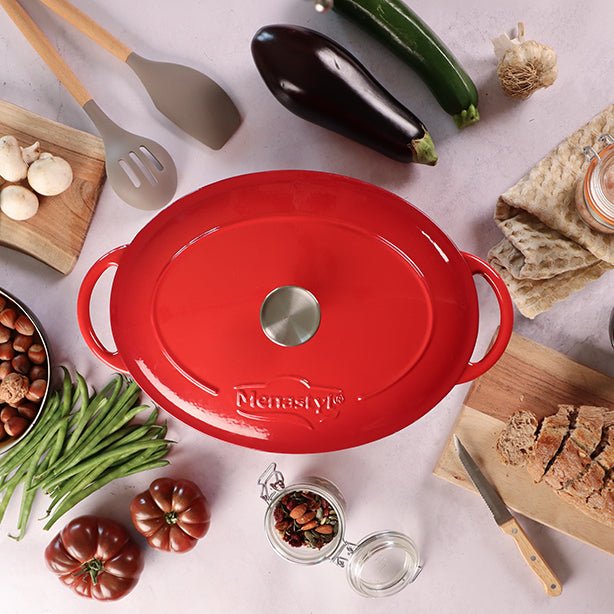Cocotte ovale Colette 9L en fonte émaillée rouge vue de dessus, entourée d'ingrédients frais et ustensiles de cuisine