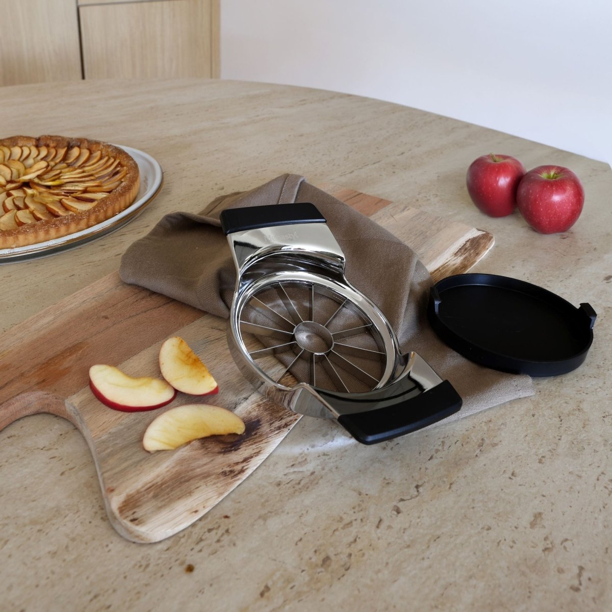 Coupe-pomme en métal avec quartiers découpés sur planche de bois, tarte aux pommes en arrière-plan