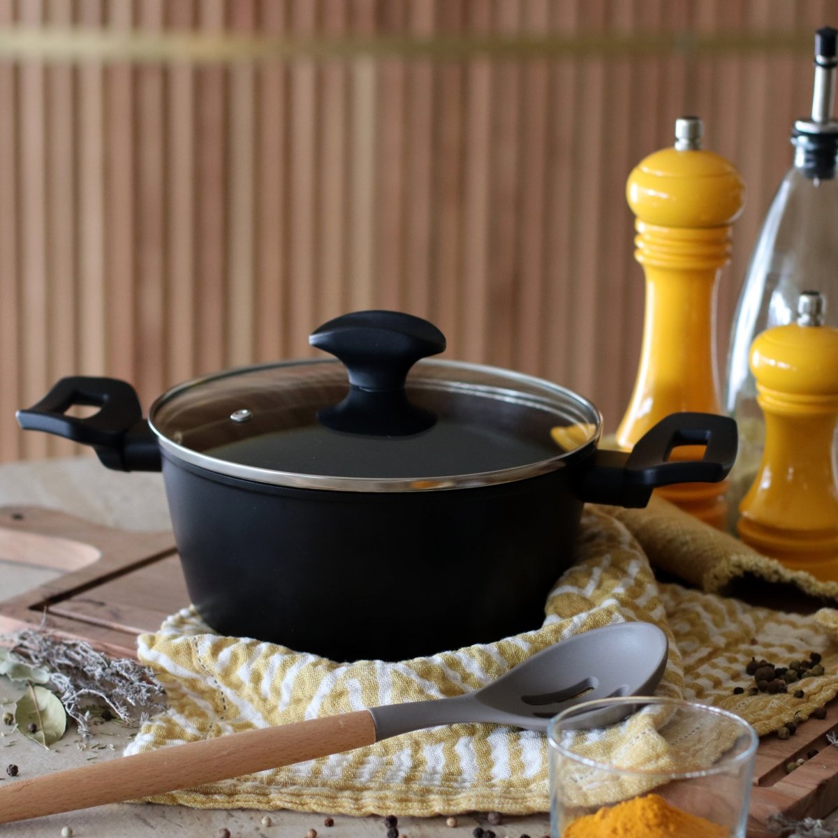 Faitout First 20 cm noir avec couvercle en verre, posé sur torchon jaune avec ustensiles de cuisine