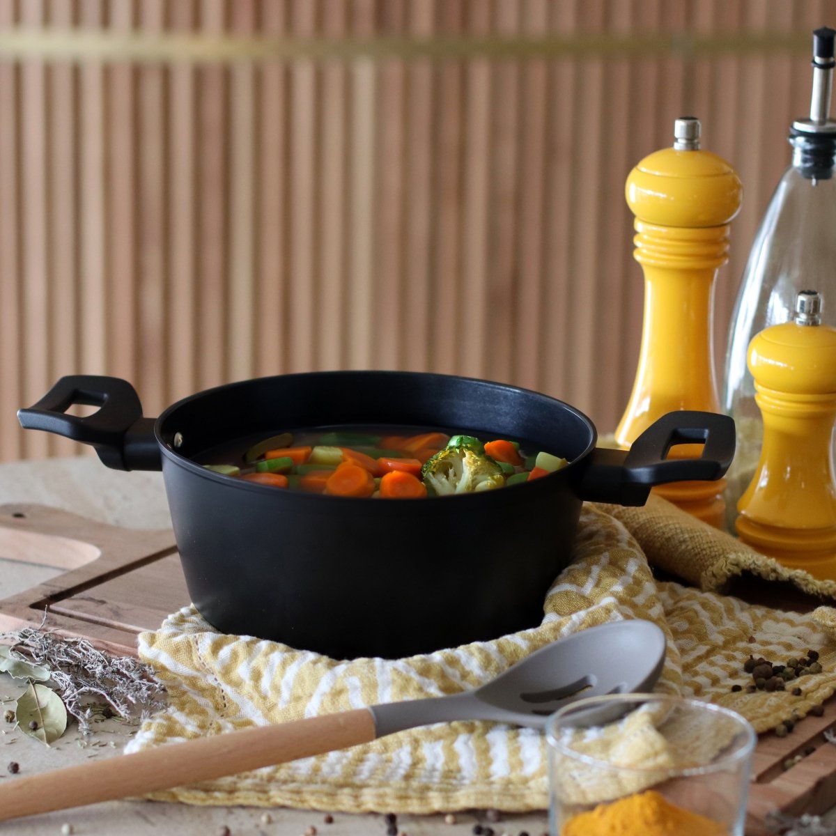 Faitout First 20 cm noir avec légumes en cuisson, posé sur tissu rayé avec assaisonnements jaunes