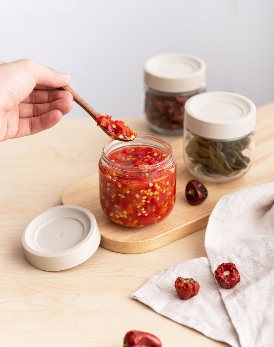 Main tenant une cuillère au-dessus d'une boîte hermétique en verre 30cl remplie de piment, sur planche en bois