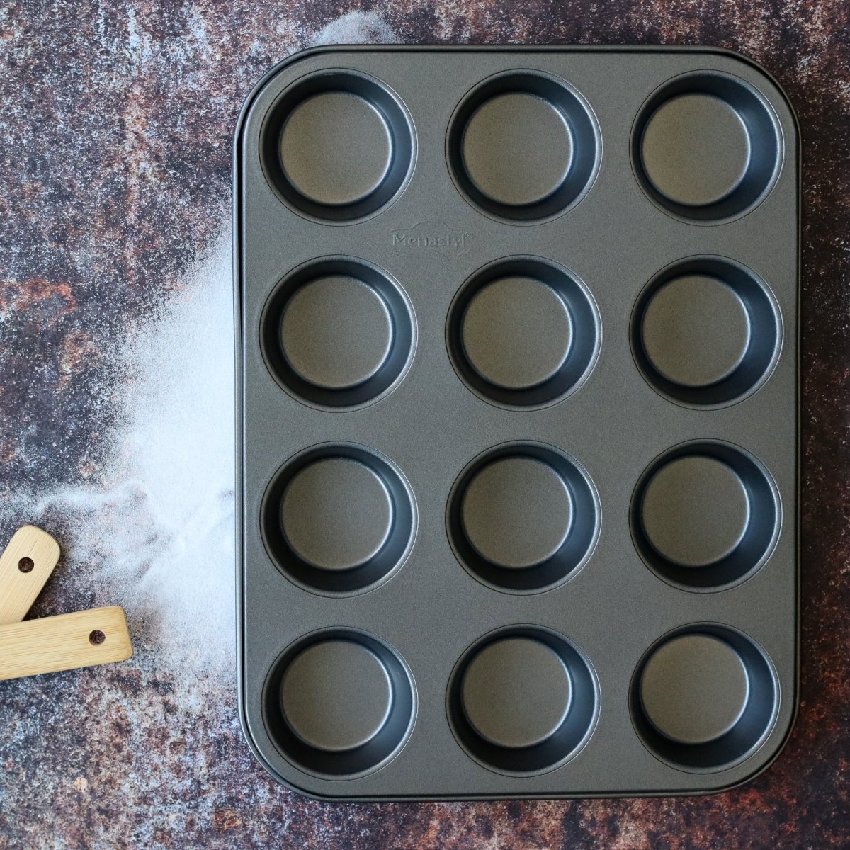 Moule à 12 muffins Amandine en acier carbone noir, vue de dessus sur surface texturée avec ustensile bois