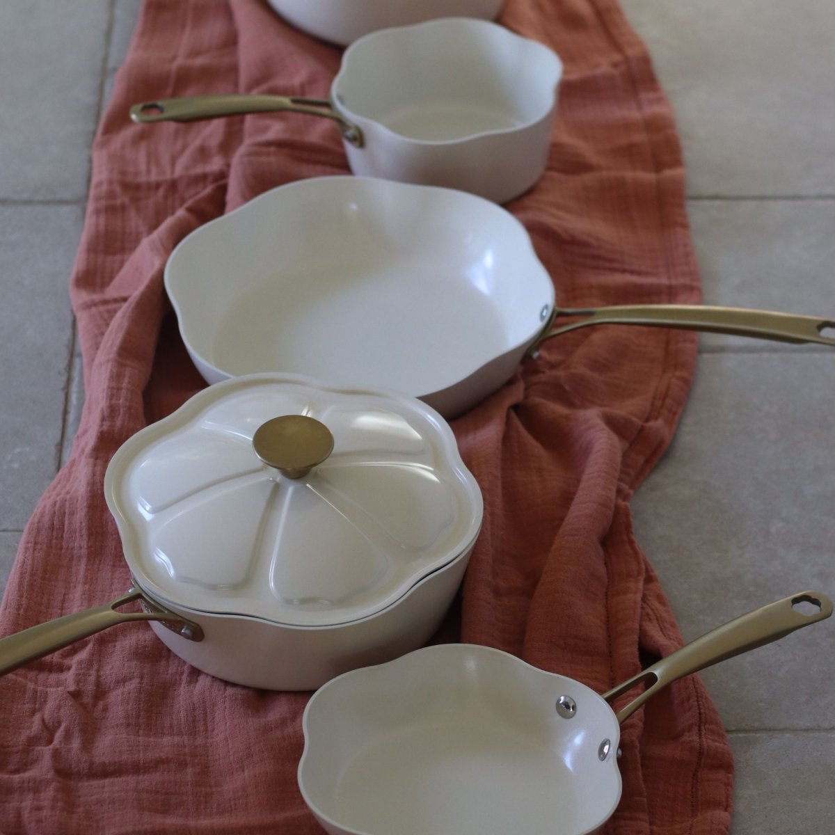 Ensemble de poêles et casseroles Dahlia beige en aluminium recyclé avec poignées dorées disposé sur tissu terracotta