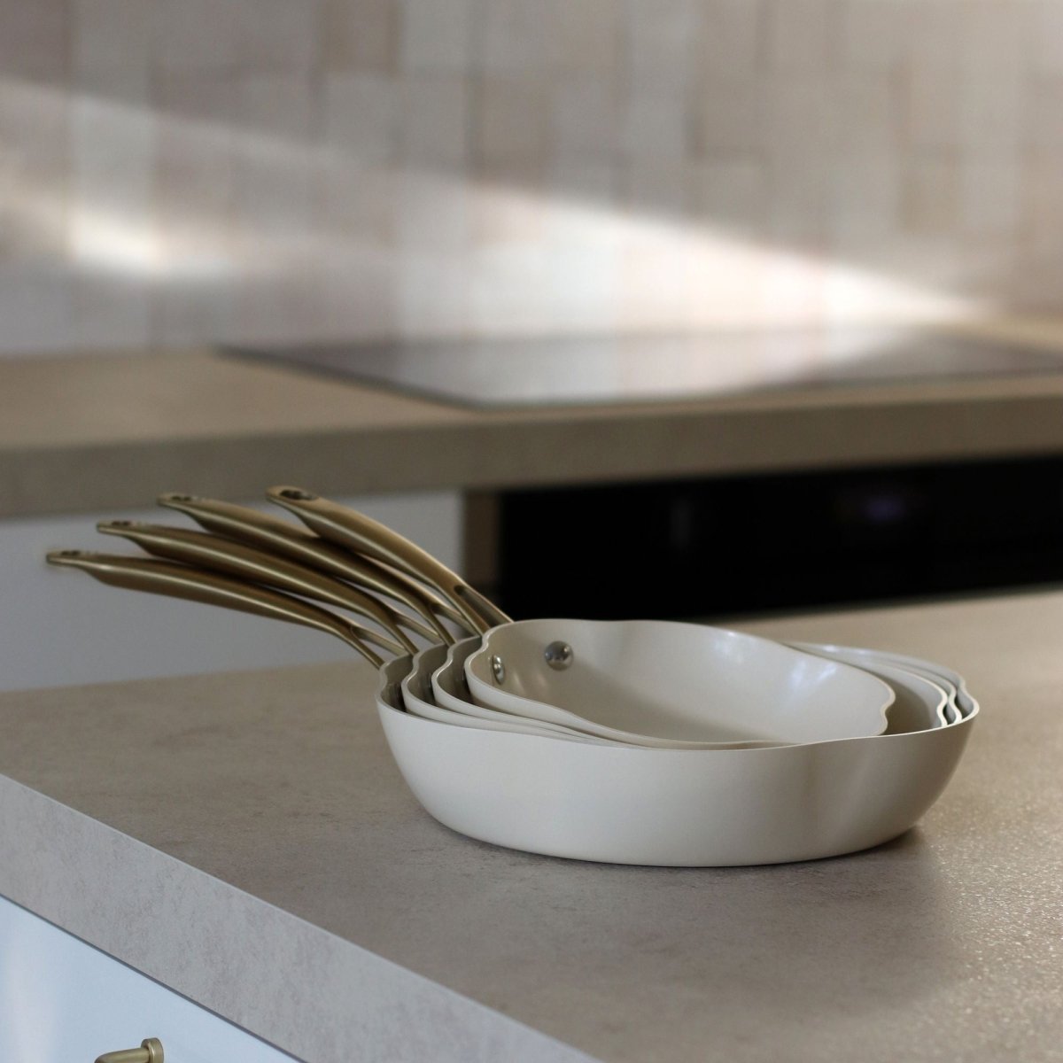 Set de 5 poêles Dahlia beige empilées sur plan de travail cuisine, vue de côté avec manches dorés