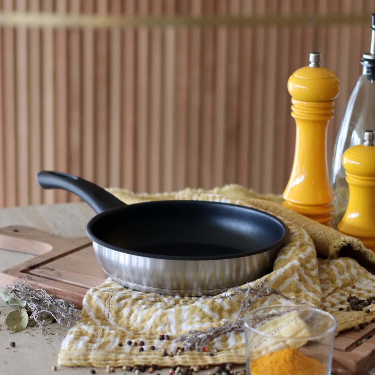 Poêle First 24 cm acier inoxydable avec revêtement antiadhésif noir, posée sur torchon jaune avec condiments