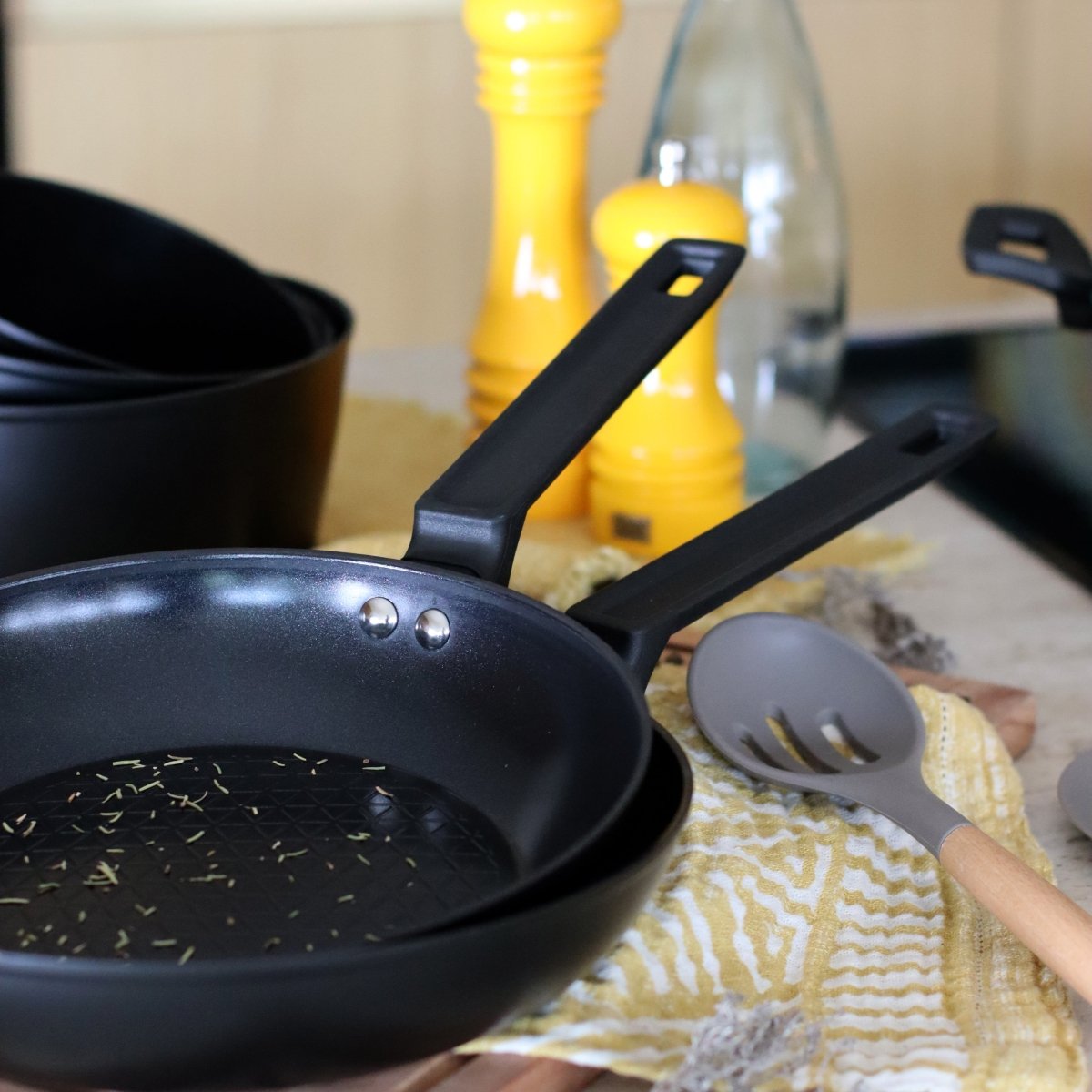 Poêle First 26 cm noire avec ustensiles de cuisine sur torchon jaune, vue d'ensemble de la gamme