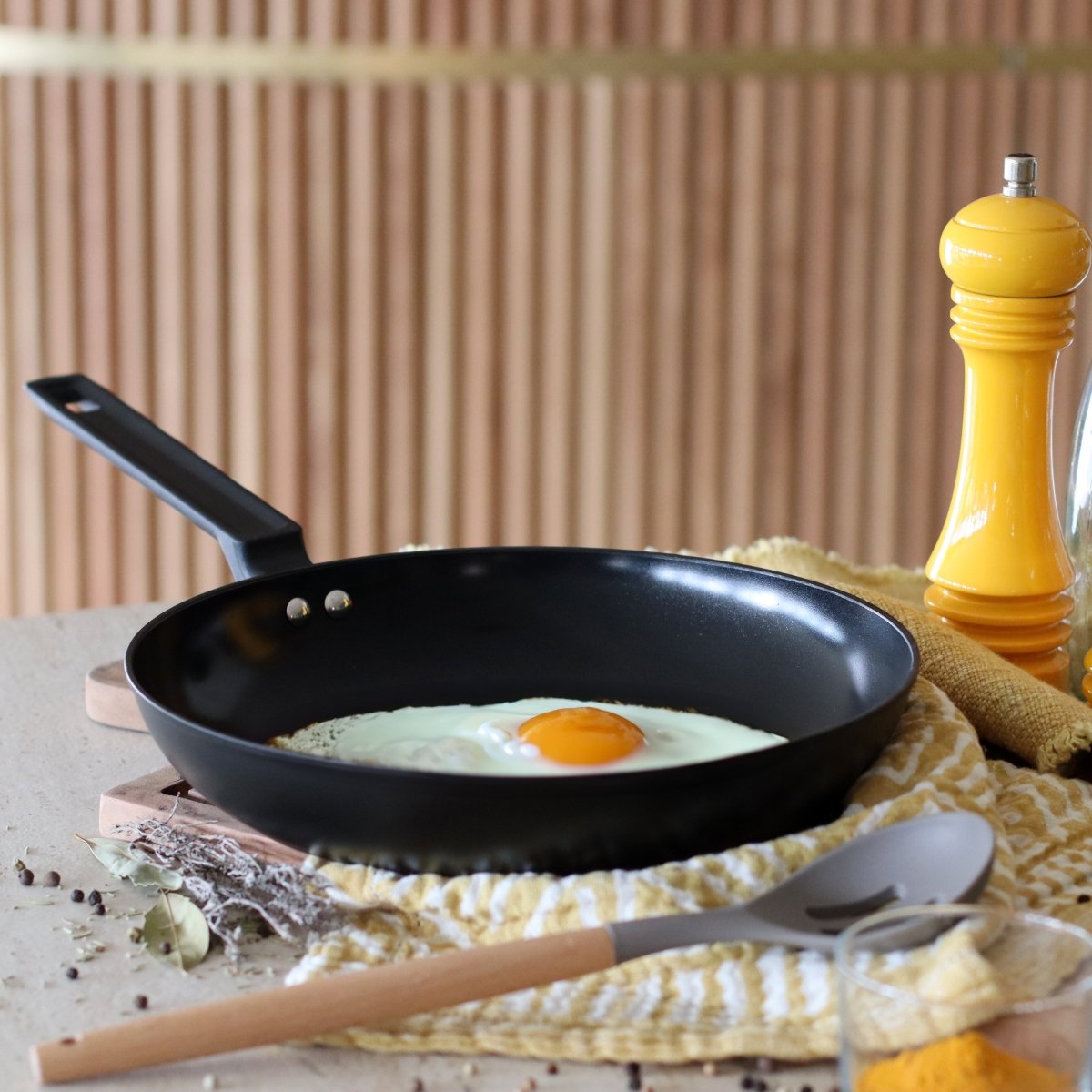 Poêle à frire First 28 cm en aluminium noir avec œuf au plat, vue d'ambiance cuisine avec moulin à poivre jaune