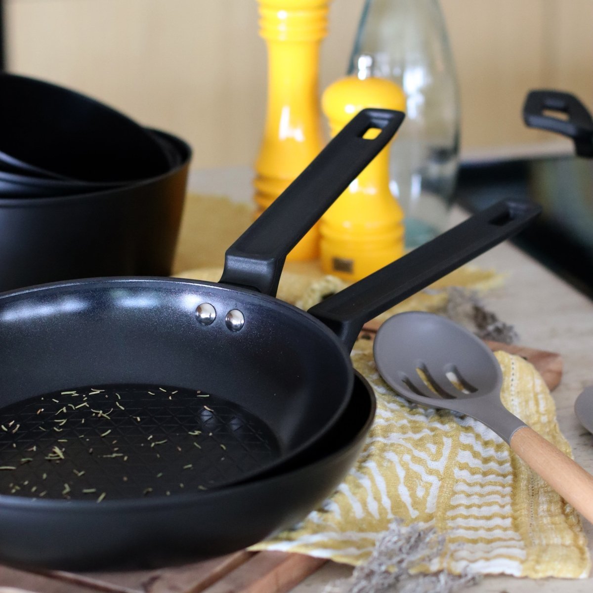 Poêle First 28cm noire avec spatule à fentes, disposée sur torchon à motifs jaunes et blancs avec ustensiles