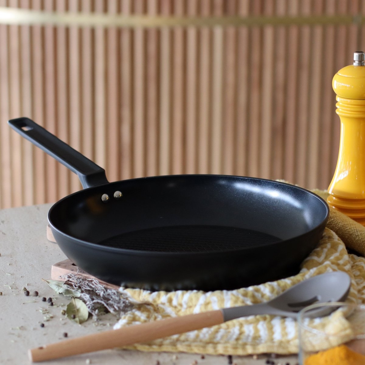 Poêle First 30 cm en aluminium noir sur plan de travail, avec ustensiles et décor cuisine