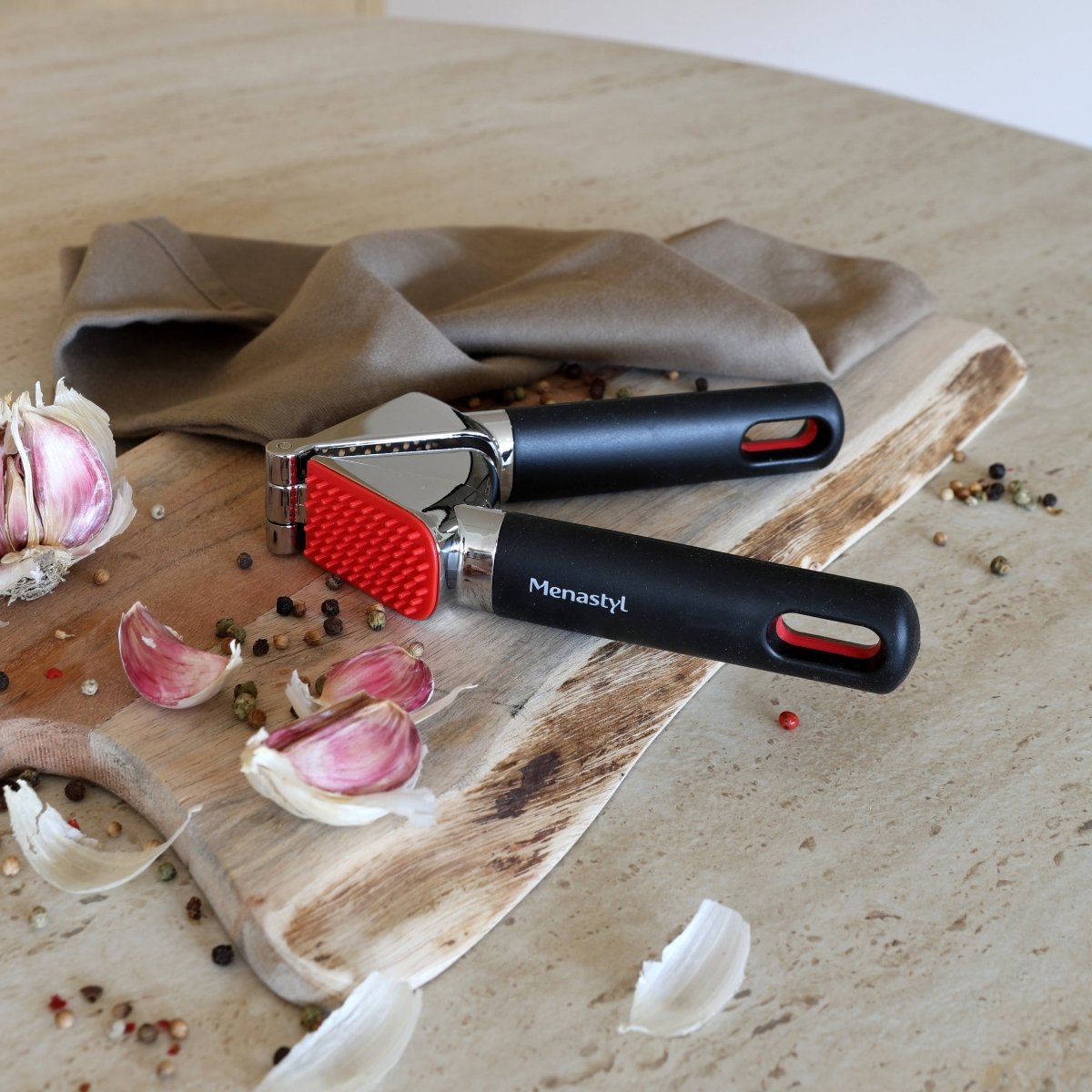 Presse-ail en acier inoxydable avec manche noir et tête rouge, posé sur planche en bois avec gousses d'ail et épices