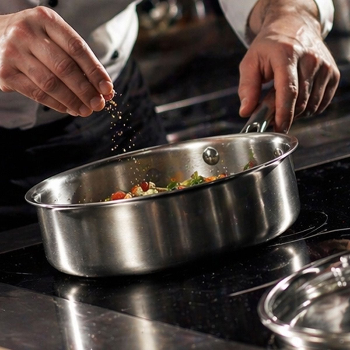 Chef en tenue professionnelle assaisonnant des légumes dans une casserole Triply en acier inoxydable sur plaque de cuisson