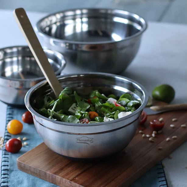 Set de 3 bols à mélanger en acier inoxydable poli, salade verte servie, posés sur planche bois avec fond clair