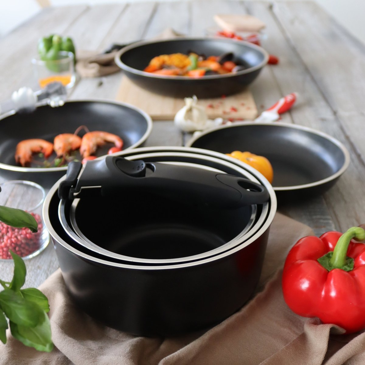 Set de 4 pièces batterie cuisine aluminium noir avec poignée amovible, casserole et poêles disposées sur table en bois avec légumes